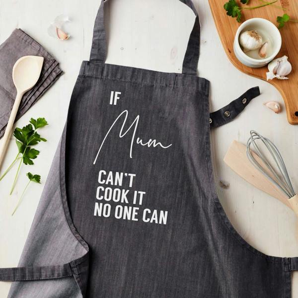 A dark grey denim apron printed with 'IF Mum CAN'T COOK IT NO ONE CAN' in white text, styled flat on a light wood surface alongside a wooden spoon, fresh herbs, and garlic.