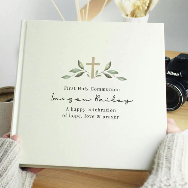 A square cream-coloured photo album featuring a golden cross motif framed by delicate green leaves, personalised with the name Imogen Bailey in an elegant flowing script.