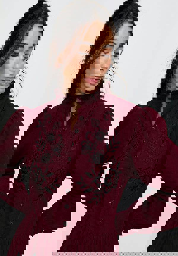 Woman wearing a burgundy dress with black floral embroidery on a plain background