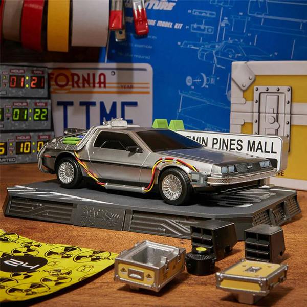 A completed silver DeLorean Time Machine model from the Back To The Future Model Advent Calendar on a display base next to miniature plutonium crates and a Twin Pines Mall sign.