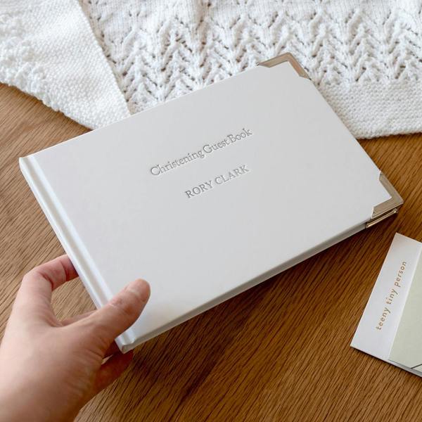 Personalised Christening Guest Book in white with silver corners, silver-foil engraved with RORY CLARK being held over a wooden table.