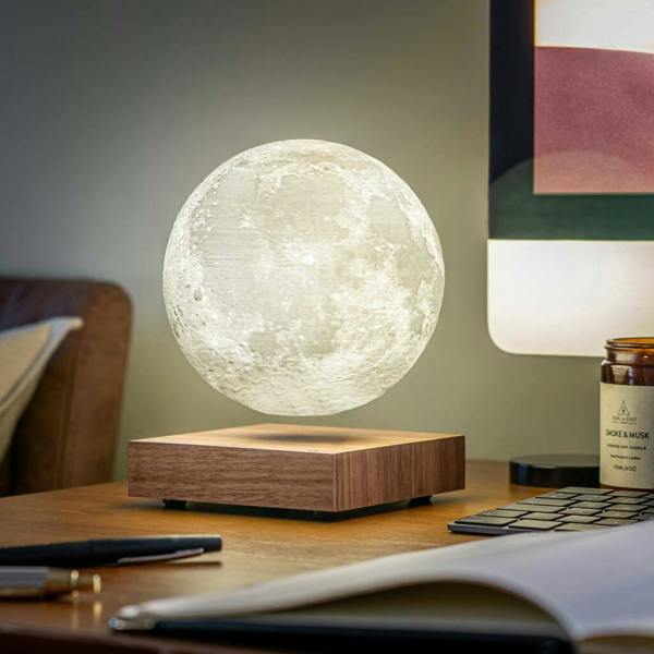 A glowing levitating moon lamp hovering above a square walnut wood base on a desk, next to a notebook and pen. The surface of the moon shows detailed crater textures.