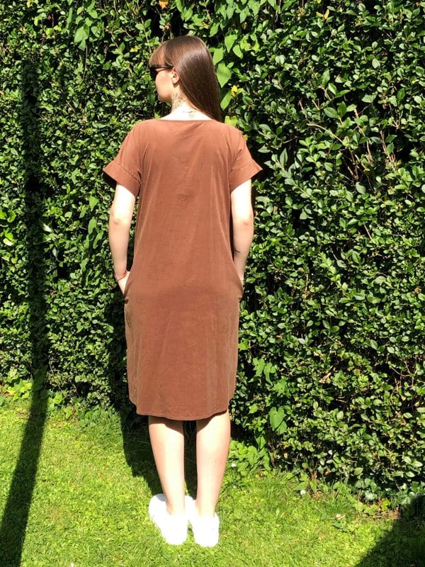 Pictured is the back of the Riverbank Dress With a Knot in Brown Corduroy. Our model is standing against a fence in the woodlands. It is a Midi length dress, worn with white trainers