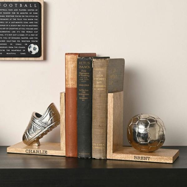 A pair of personalised wooden football bookends with engraved bases reading Charlie and Brent, holding books on a dark desk beneath a football sign.