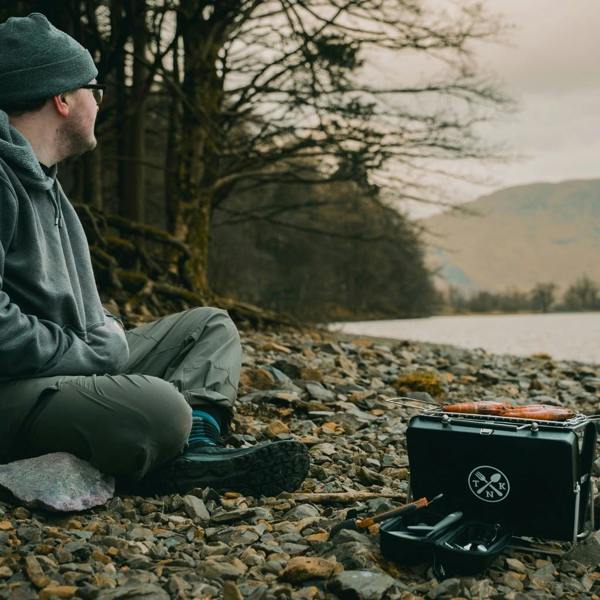 Man sitting on a rocky shore next to a Portable Notebook BBQ with a view of mountains.