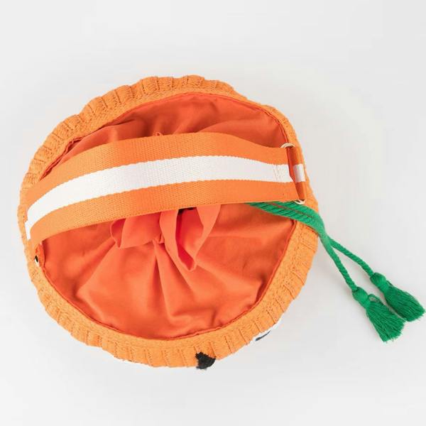A top-down studio view of an orange woven pumpkin trick-or-treat basket bag, showcasing the gathered orange polycotton interior lining, the striped webbing handle, and the green tassels.