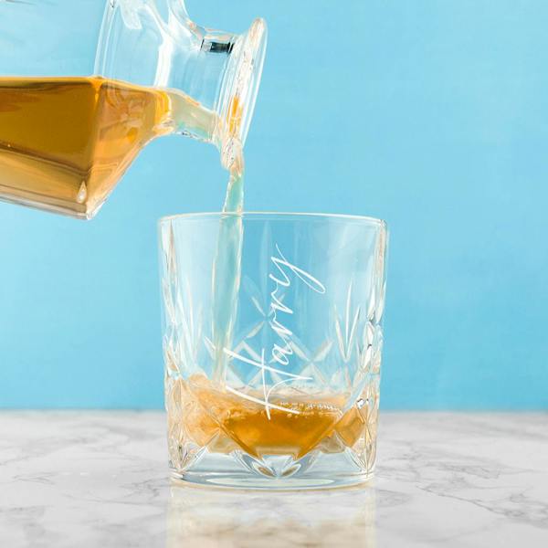 An intricate crystal whisky tumbler engraved with the name 'Harry' being filled with golden liquor from a matching glass decanter on a marble surface.