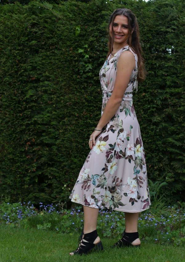 This is a young lady wearing a floral printed dress in pale pink. It has a mock wrap style V neck style top, which has gathers under the bust and the shoulders with a tie on the front. The model is wearing black lace up heels, she is standing to the side and looking towards the camera, silhouetted against a lush garden.