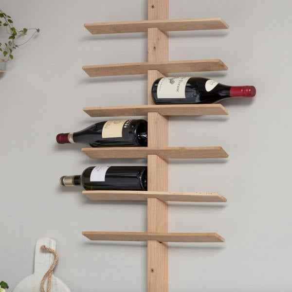 A tall wall-mounted raw oak wine rack holding multiple bottles of red wine horizontally on left and right staggered wooden slats against a light grey wall.