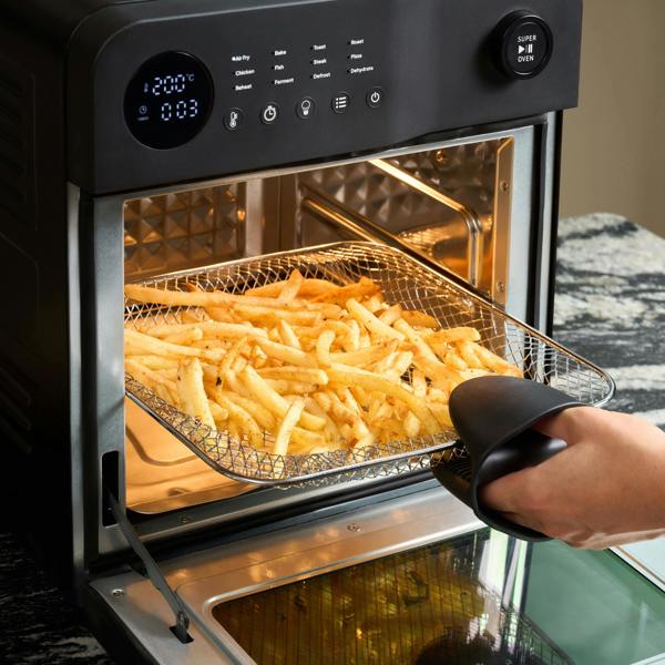 A hand in an oven mitt removes cooked fries from a Fridja f66 Super Oven - 15L Digital Air Fryer Oven, which features a PFAS free design and a digital display showing temperature and time for precise convection cooking.