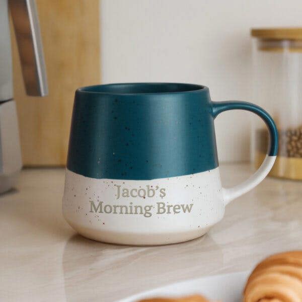 A two-tone blue and speckled white ceramic mug printed with 'Jacob's Morning Brew' sitting on a kitchen counter next to a croissant.