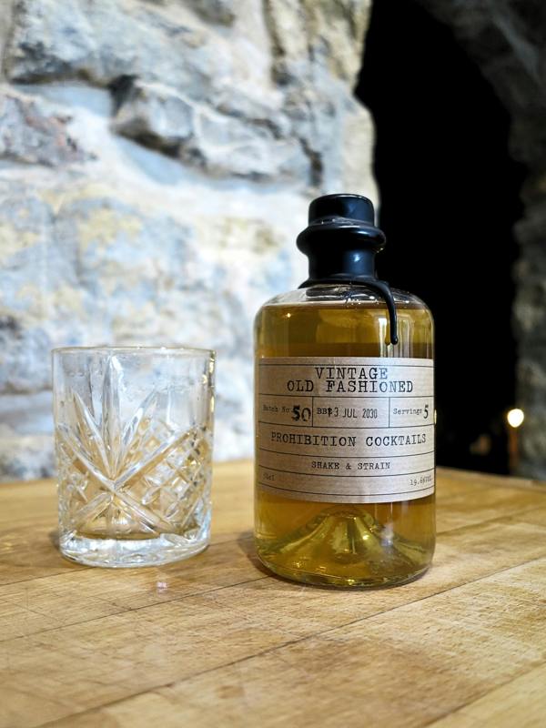 A bottle of 'Vintage Old Fashioned' prohibition cocktails with amber liquid, next to a cut-crystal tumbler, set on a wooden table with a rustic stone wall background.