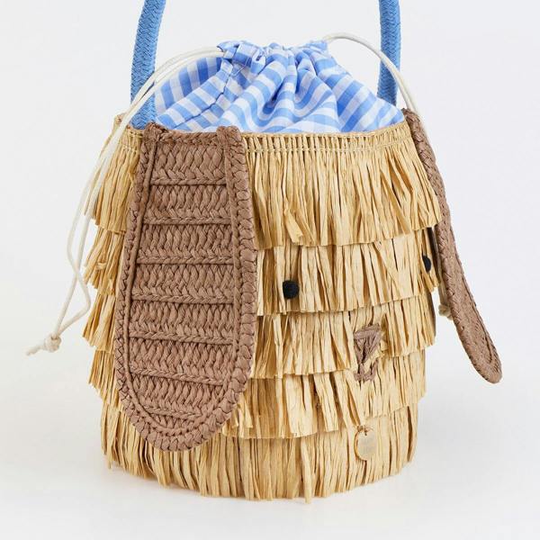 A close-up side angle of the woven raffia puppy basket bag showing the texture of the floppy brown ear and tiered straw fringe.