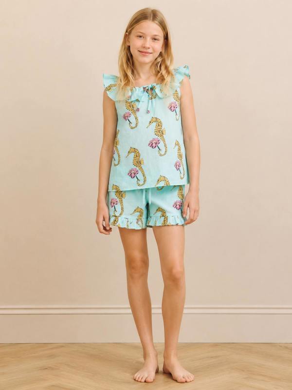 A young girl stands barefoot against a beige wall and wood floor, wearing a two-piece pajama set in light aqua with a seahorse print. The set includes a ruffled sleeveless top with a tie at the collar and matching shorts.