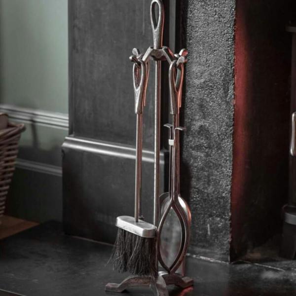 A traditional dark pewter cast iron fireside companion tool set standing on a dark hearth next to an open fireplace.