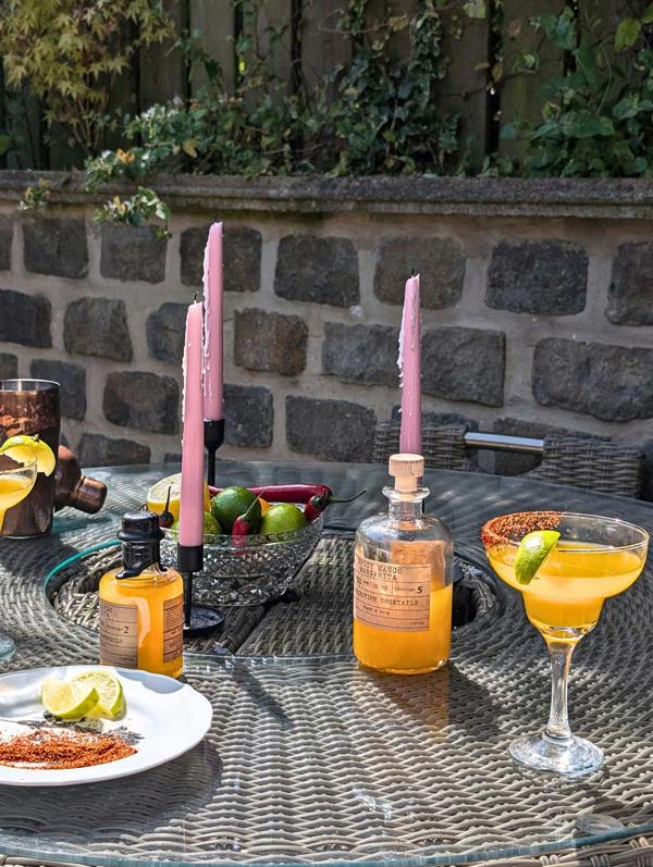 Outdoor patio table setup with pink taper candles, a glass of yellow cocktail, spice plates, a bottle of orange liquid, and a small bottle on a woven wicker table in front of a stone wall.