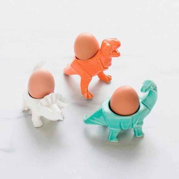Three ceramic dinosaur-shaped egg cups—a triceratops, T-rex, and brachiosaurus—each holding a boiled egg, on a clean white surface.