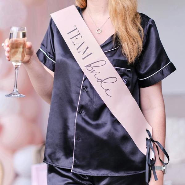 Close mid-section view of a bridesmaid wearing a pale pink cursive script sash over a black satin pyjama set while holding a champagne glass.