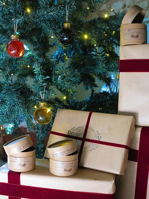 Three gift sets from Prohibition Cocktails with cream-colored labels and lids are arranged on and beside wrapped gift boxes tied with red ribbons, set in front of a decorated Christmas tree with warm string lights.