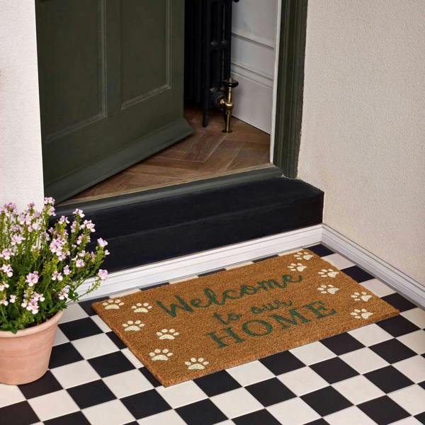 Doormat with 'Welcome to our home' text on a checkered floor, next to a potted plant.