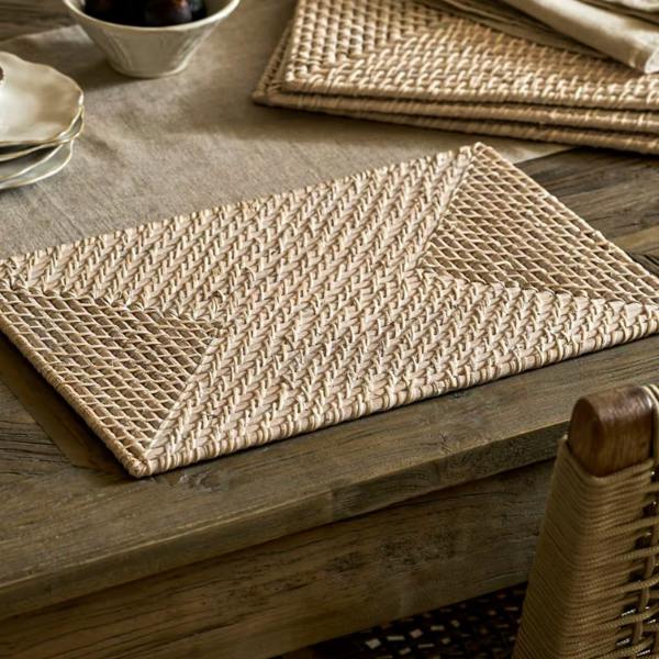 Handwoven white washed rectangular rattan placemat styled on a rustic wooden dining table alongside silverware and napkins.