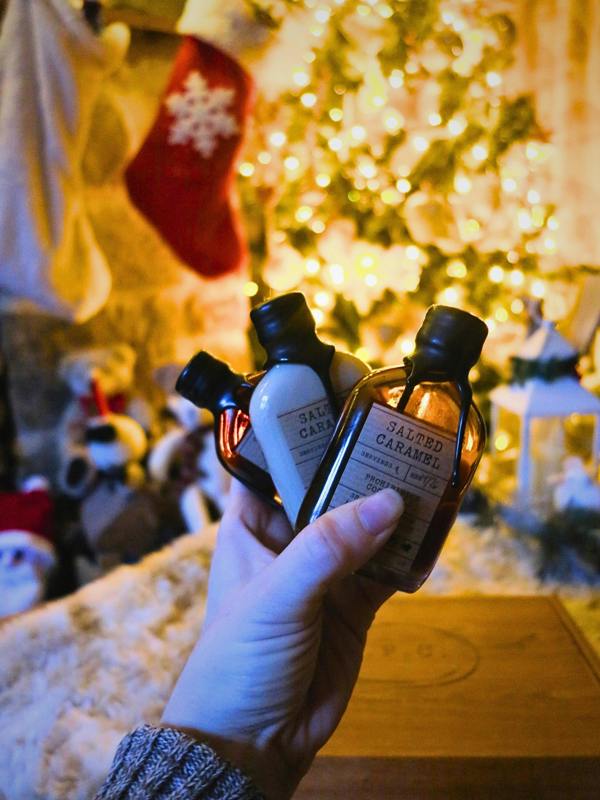 A hand holds three small cosmetic bottles in front of a warm, festive background with a glowing Christmas tree and stockings. The bottles are Salted Caramel