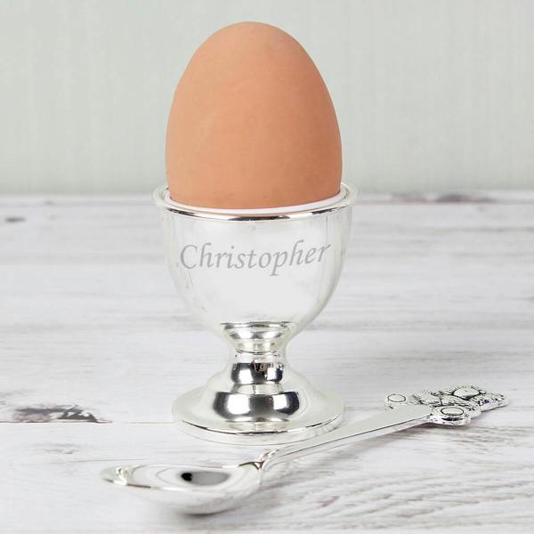 A polished silver-plated egg cup engraved with the name "Christopher" holding a brown egg, alongside a matching ornate silver spoon, resting on a white wooden surface.