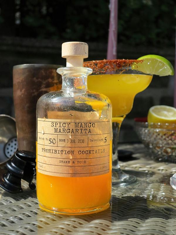 A clear glass bottle labeled 'Spicy Mango Margarita' filled with bright orange liquid, placed on a woven table in an outdoor setting with a yellow margarita glass and lime garnish behind it.