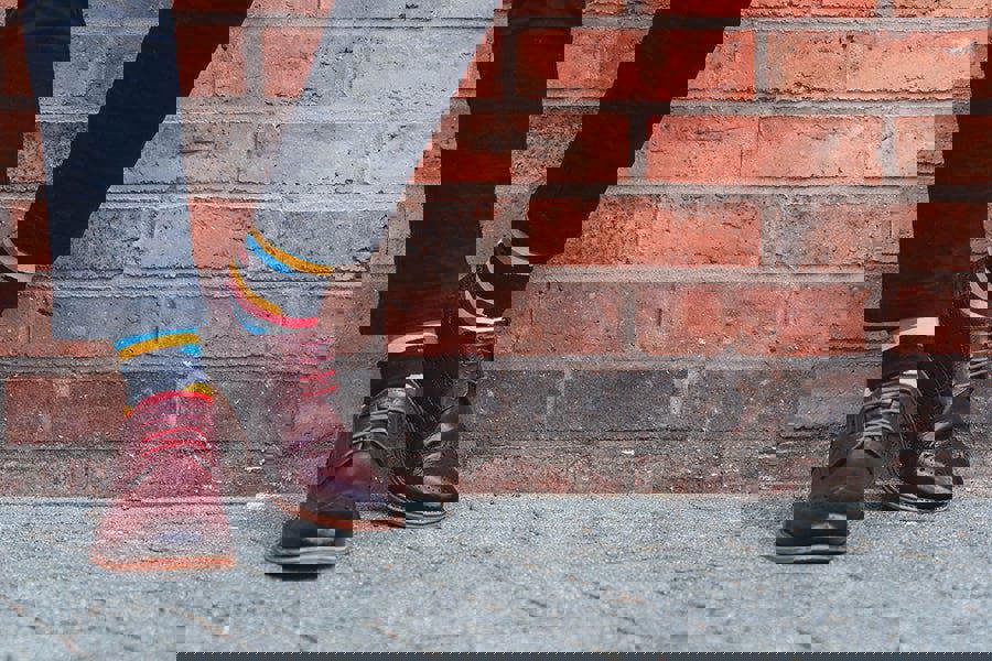 Person wearing burgundy shoes and colourful crew socks standing against a brick wall.