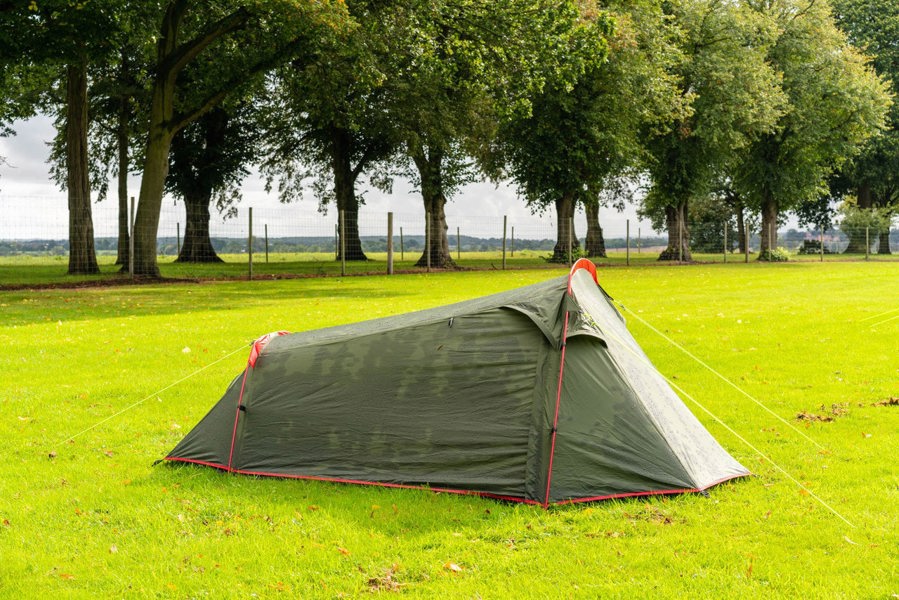 The OLPRO Beckford Lightweight 2 Person Tent pitched in a field with a treeline behind it with its doors closed, depicting a wild camping scene.