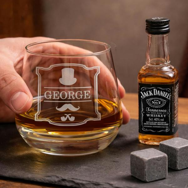 A hand holding an engraved Personalised Gentlemen Tumbler filled with whiskey next to a 50ml miniature Jack Daniel's bottle and slate whisky stones.