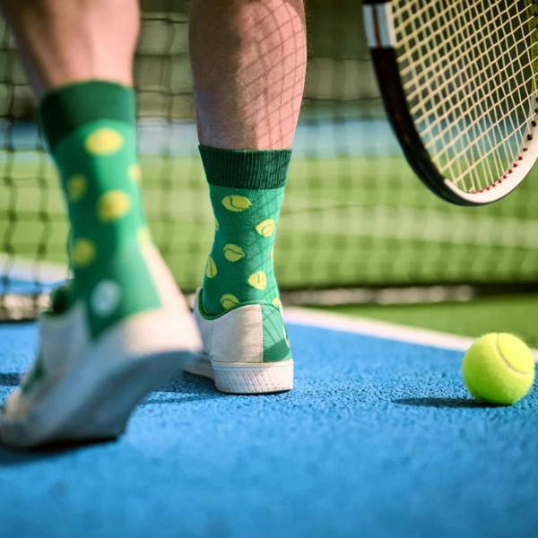Low-angle shot of a person on a blue tennis court wearing green socks with yellow tennis balls pattern next to a tennis racket.
