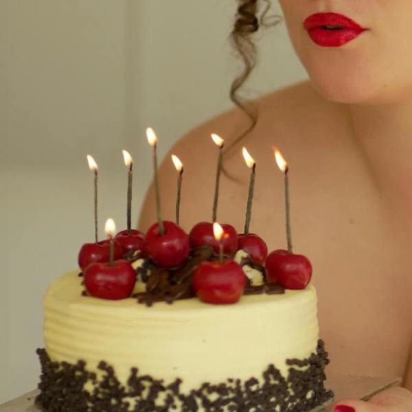 A close-up lifestyle shot of a person with red lipstick holding a small frosted birthday cake topped with eight lit, realistic cherry-shaped candles.