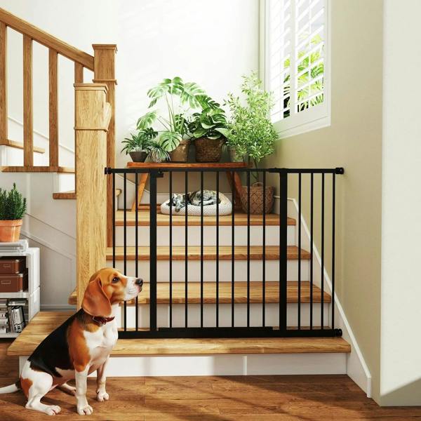 Black stair safety gate with vertical bars installed in a hallway, dog sitting nearby