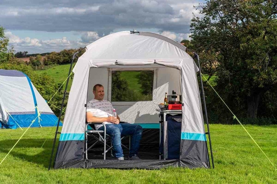 The OLPRO Pod Kitchen/Storage Tent pitched on a campsite with its door open with a male sat inside with a camping stove and a tree line in the background.