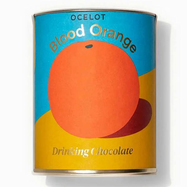A contemporary cylindrical product tin with a bold blue and yellow color-blocked background featuring a large orange graphic and 'Blood Orange Drinking Chocolate' text.
