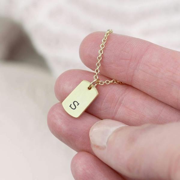 A dainty gold-plated rectangular tag pendant necklace engraved with a custom initial, resting on a clean white surface.
