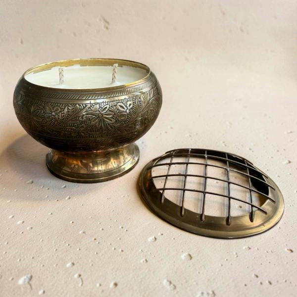 An ornate brass scented candle in a decorative bowl-shaped container with floral engravings, shown with its matching metal lid placed beside it on a textured beige surface.