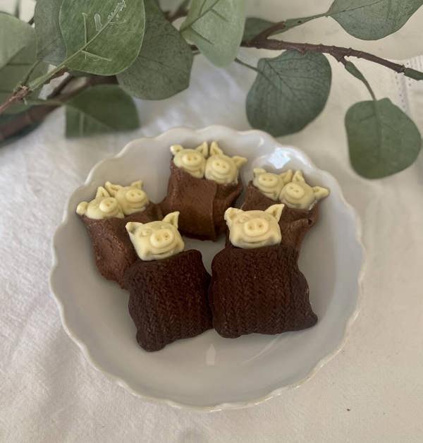 Six novelty Belgian chocolate pigs in blankets in a white scalloped bowl against a festive botanical background.