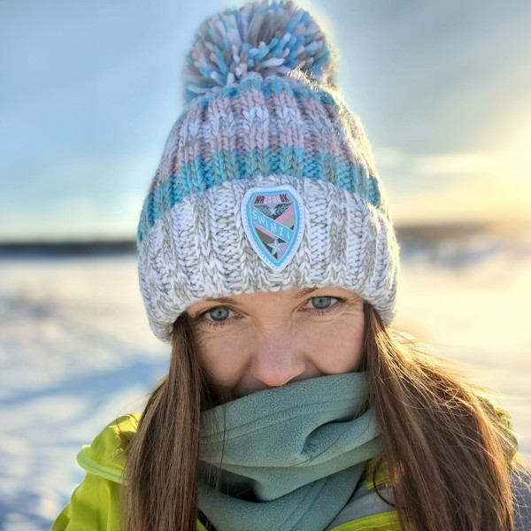 Set against a snowy arctic landscape woman is wearing a white and light grey based super bobble hat with cool pastel pink and blue stripes.  The hat has a deep ribbed headband, contains reflective yarns and is lined in a luxurious sherpa fleece. 