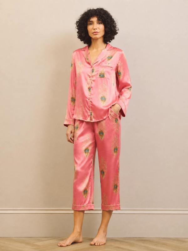 A woman modeling a two-piece satin pajama set in pink with a green and gold floral pattern, standing against a neutral beige wall.