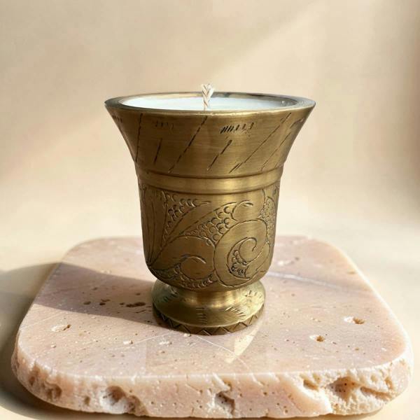 A brass candle holder with engraved floral patterns, holding a white candle, displayed on a square beige stone slab against a warm beige background.