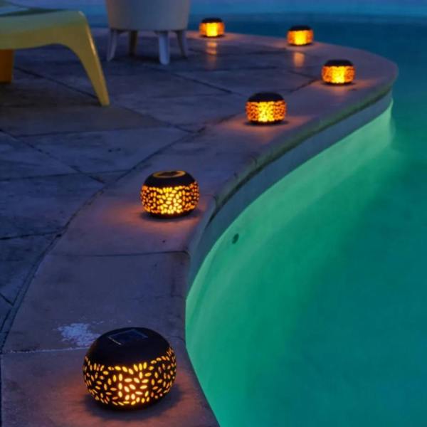 A row of glowing Moroccan solar powered filigree flame lanterns reflecting warm amber light along the edge of a swimming pool at night.