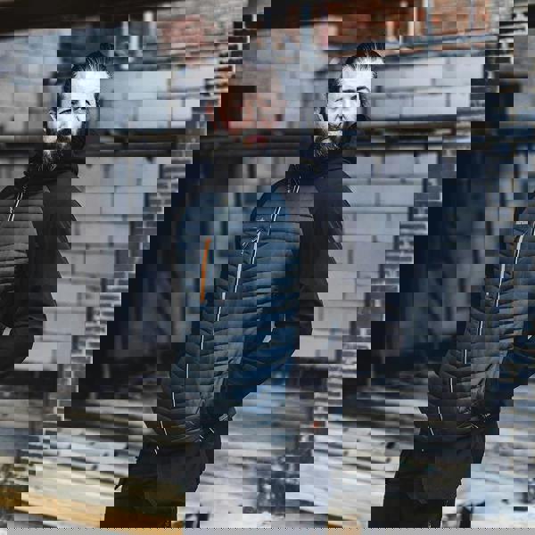 Scruffs Mens Trade Thermo Jacket Zip Up Work Warm Hooded Jacket Coat, a man wearing a black and dark grey insulated work jacket with a hood, standing outdoors in an industrial setting.