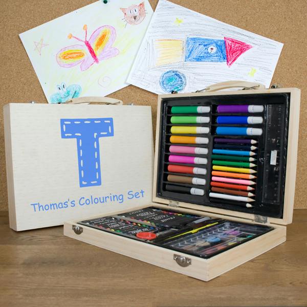 A 63-piece personalised children's colouring set in a wooden case printed with a large blue letter T and Thomas's Colouring Set, open on a desk displaying art supplies alongside drawing artwork.
