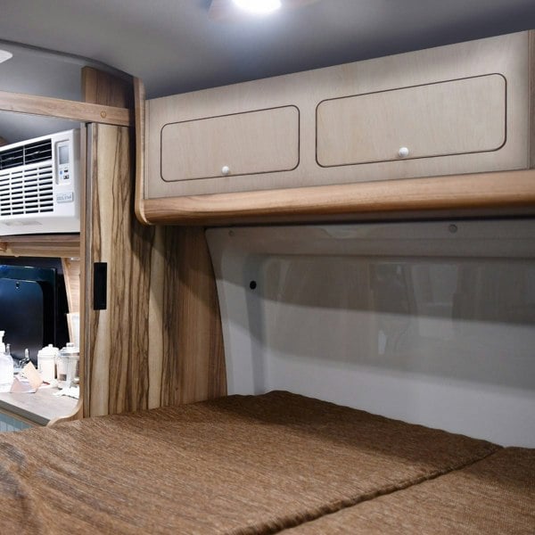 Overhead camper cabinet in light wood with two drawers, mounted above a cozy sleeping area, showcasing a sleek, modern design