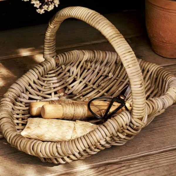 A hand-woven natural rattan trug basket containing wooden-handled garden tools and twine sitting on a rustic wooden floor next to a terracotta pot.