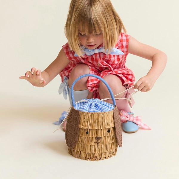 A child in a red gingham dress adjusting the blue drawstring lining of a woven raffia puppy basket bag with floppy brown ears.