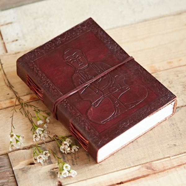 The Sitting Buddha Leather Journal, a closed brown leather piece embossed with its namesake design and secured by a strap, rests gracefully on a wooden surface with small white flowers artfully arranged beside it.