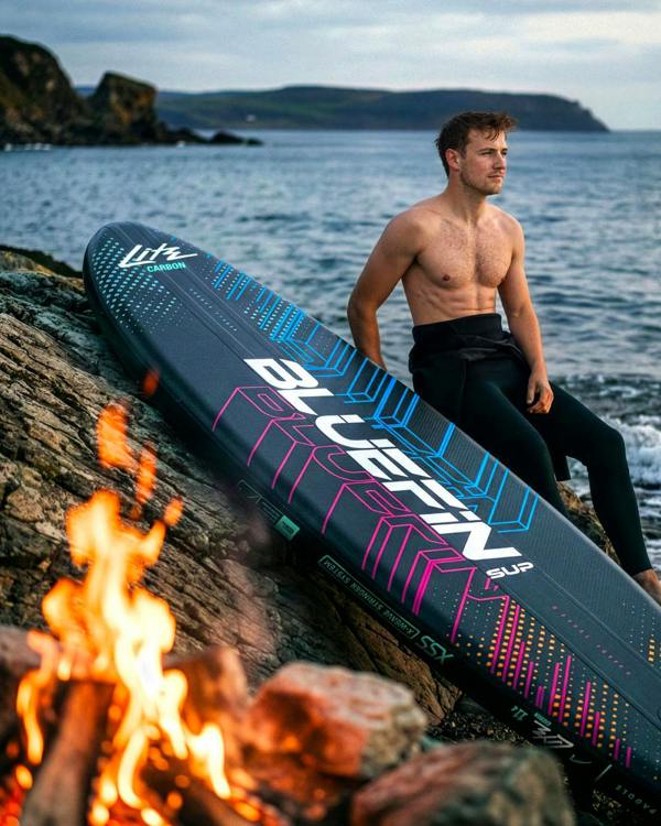 Man sitting on rocks by the sea with a Bluefin SUP board and a campfire in the foreground.
